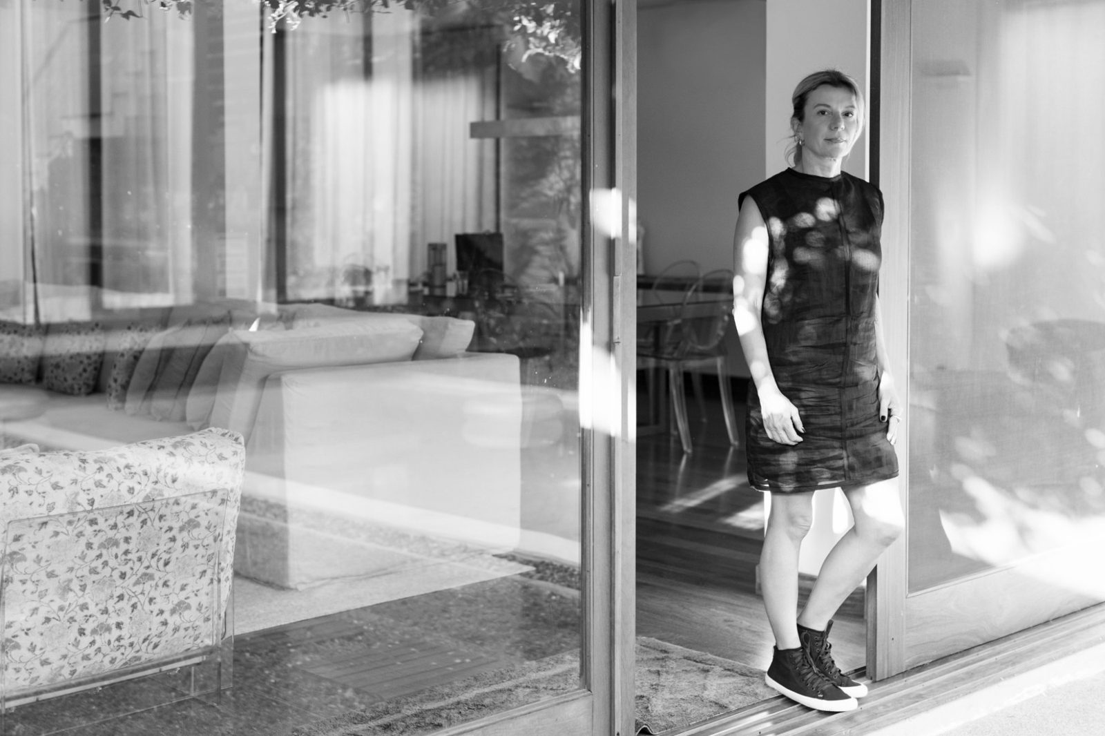 Fernanda Cerávolo de vestido preto, encostada numa porta de vidro de casa, em foto preto e branco