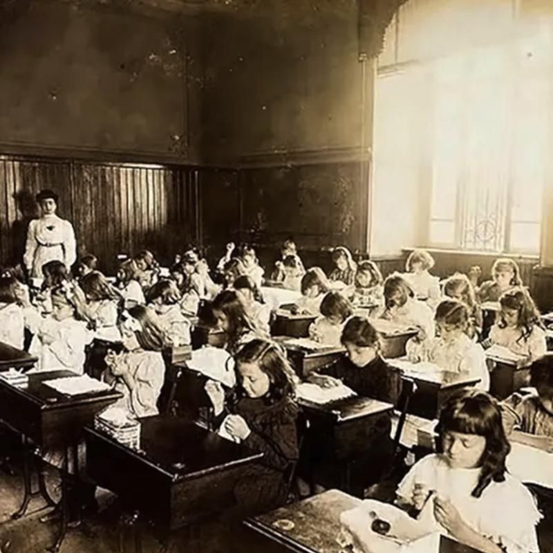 Sala de aula antiga cheia de meninas em carteiras, com uma professora ao fundo